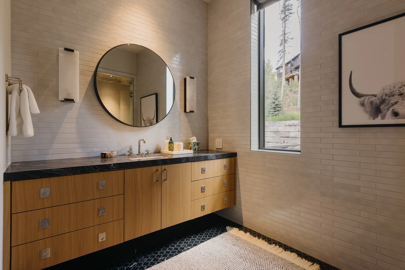 A bright bathroom with a wood vanity, round mirror, and large window.