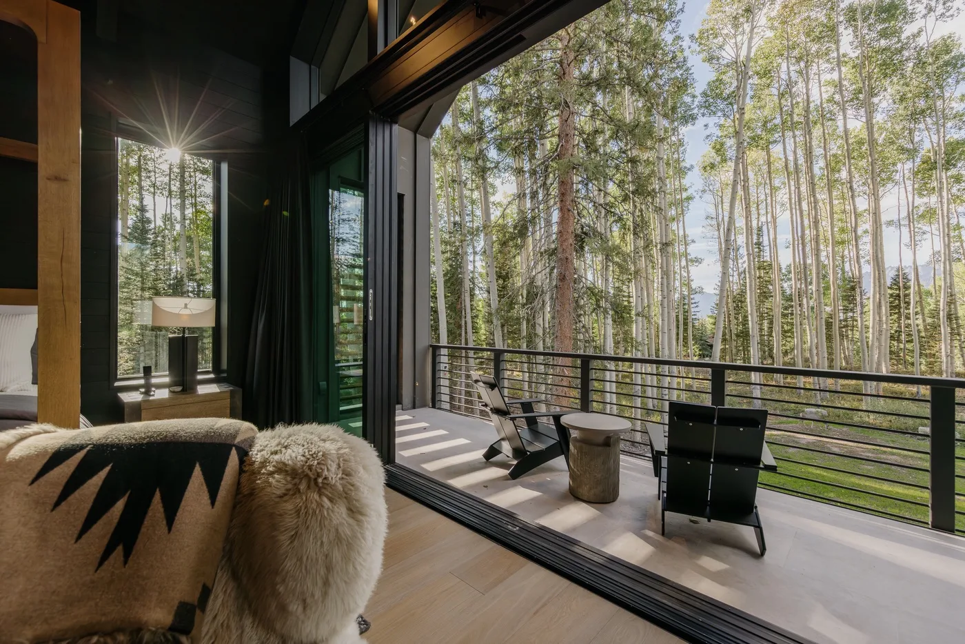 A bedroom opens onto a balcony with seating and a view of tall trees.