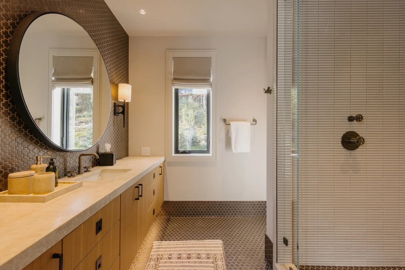 A bright bathroom has a long vanity, large round mirror, and glass shower.