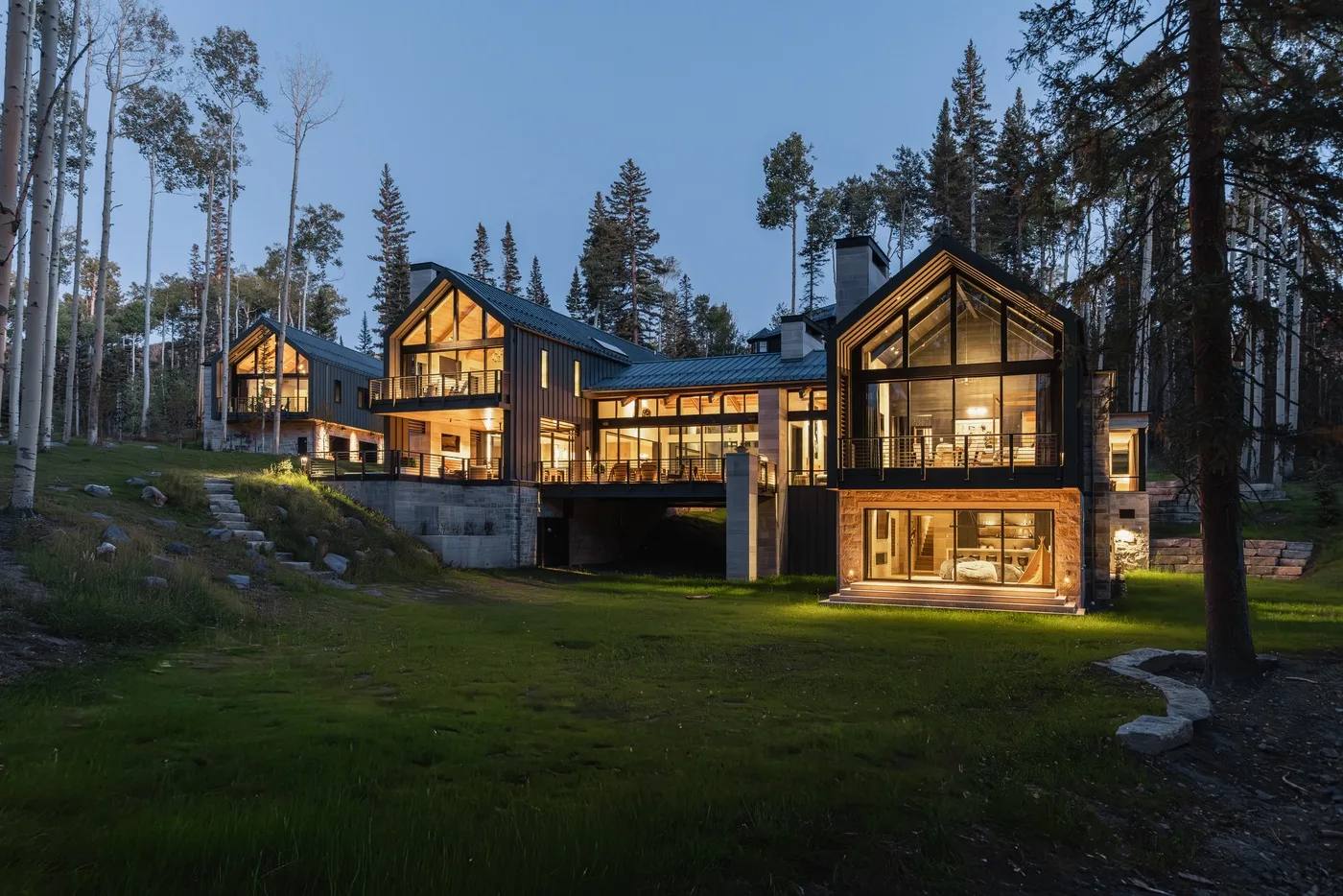 A modern house with large illuminated windows stands among tall trees.
