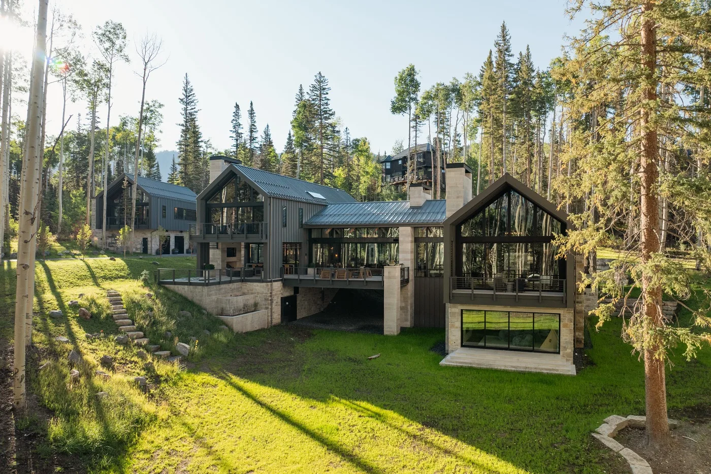 A large modern house sits on a grassy slope among tall trees.