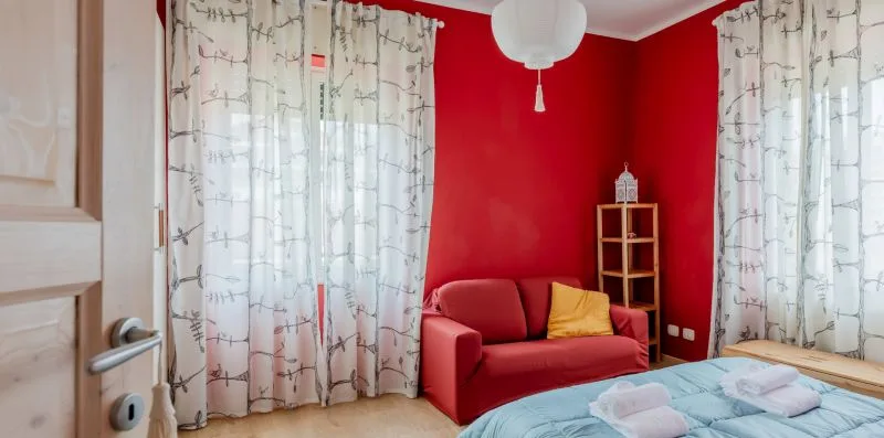 Un dormitorio con una pared roja, una zona de estar y una cama junto a ventanas con cortinas.