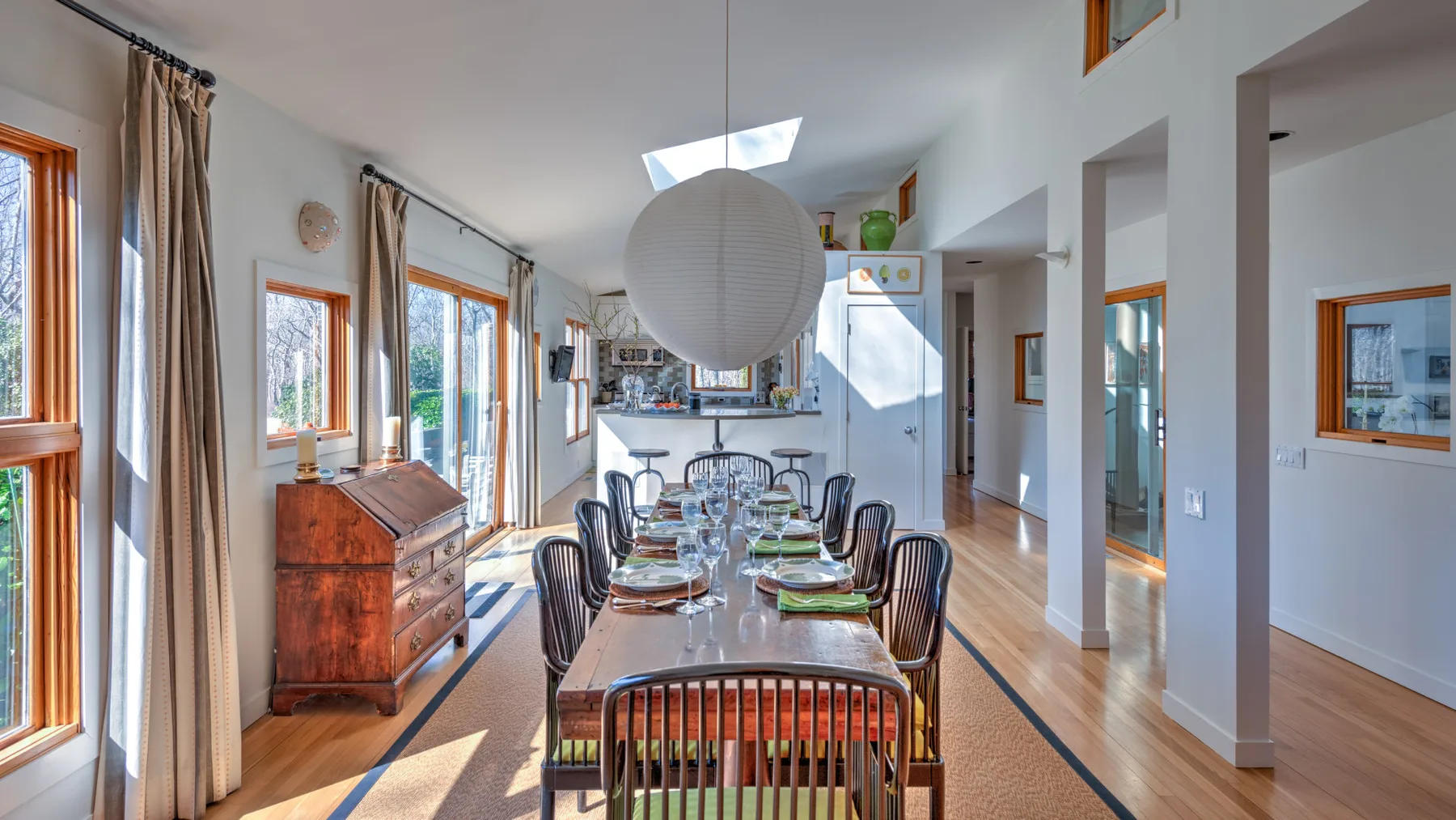 A bright dining area has a long table set beside tall windows.