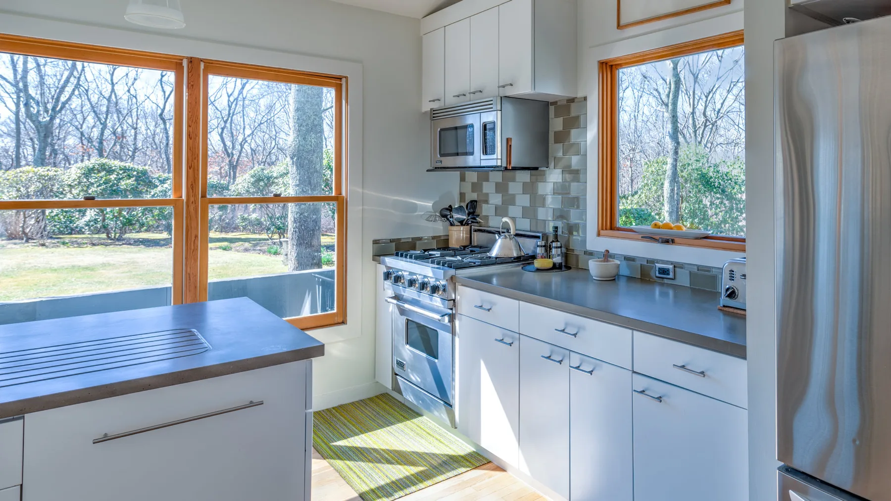 Una cocina luminosa con gabinetes blancos y grandes ventanas con vista a los árboles.