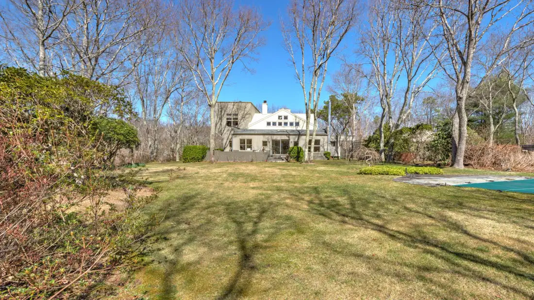 A grassy yard with mature trees, shrubs, and a house in the background.