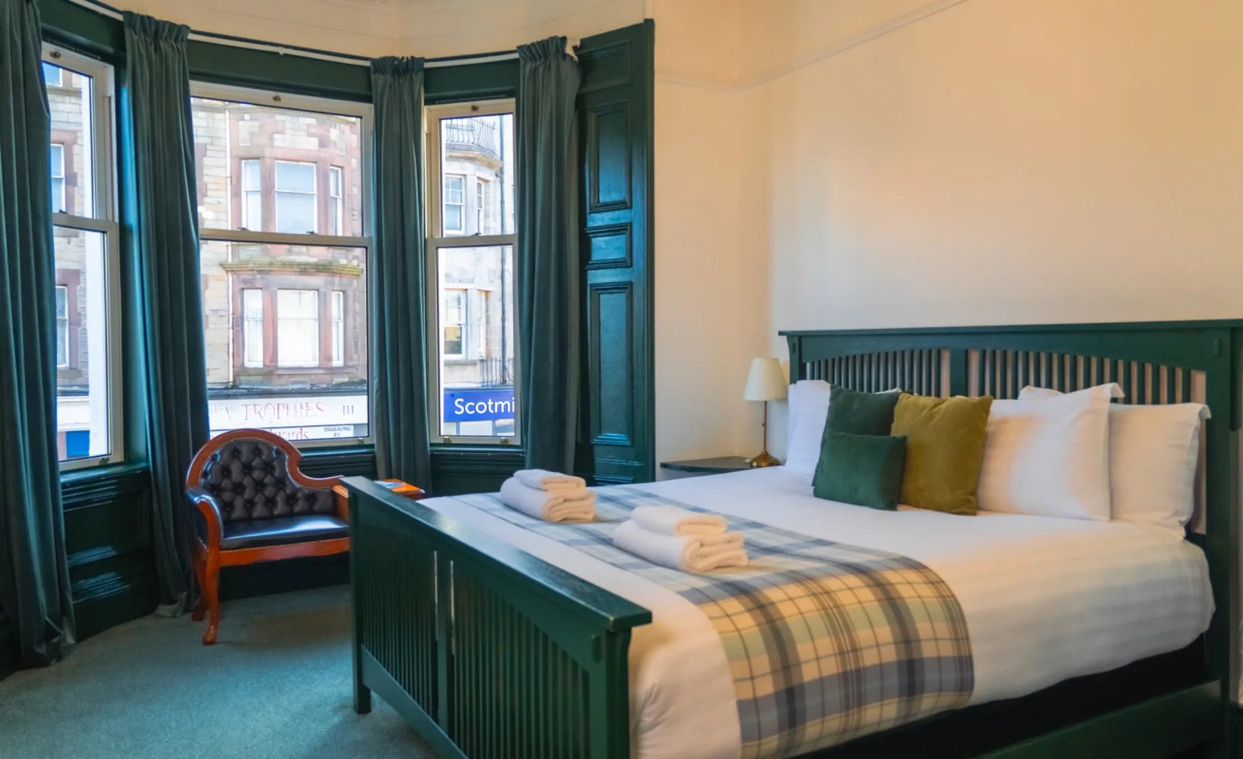 Dormitorio con una cama doble, una silla y grandes ventanas con cortinas verdes.