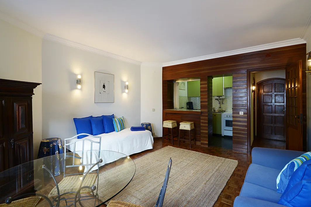 A bright studio living area with a white daybed, glass table, and blue seating.