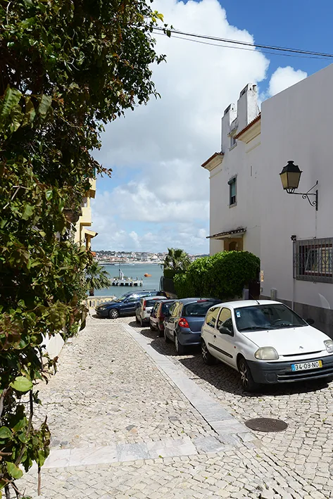 Una calle empedrada con coches aparcados, árboles y el paseo marítimo al fondo.