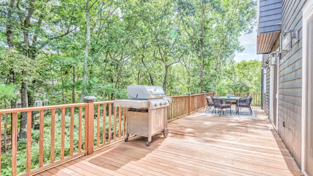 Una terraza de madera con una parrilla y comedor exterior da a una zona arbolada.