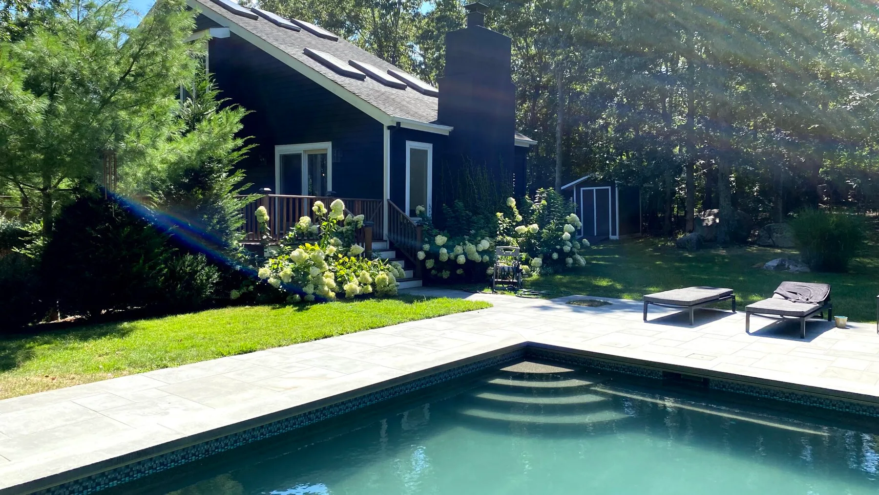 Una piscina en el patio junto a una casa oscura, con asientos en la terraza pavimentada.