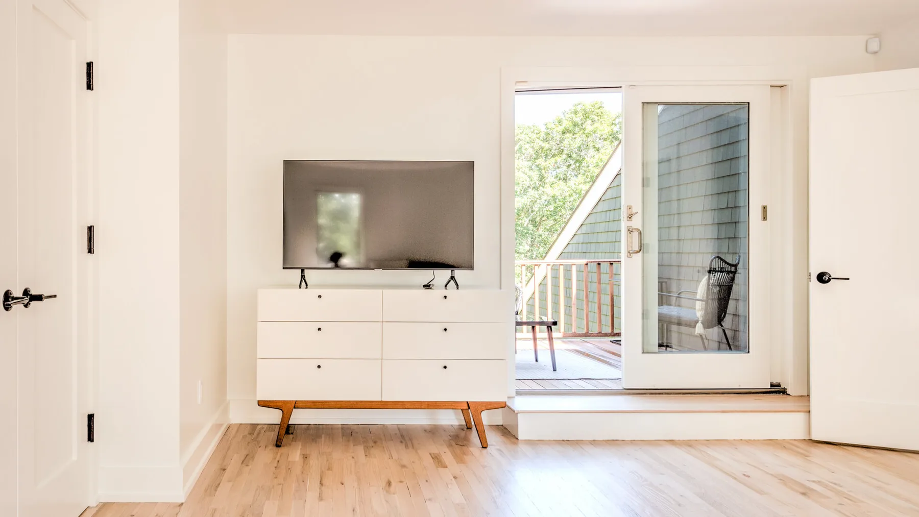 Un dormitorio luminoso con un televisor sobre una cómoda junto a una puerta abierta al balcón.