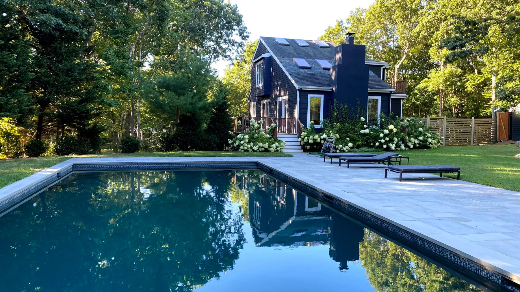 Una piscina y un patio pavimentado están junto a una casa oscura rodeada de árboles.