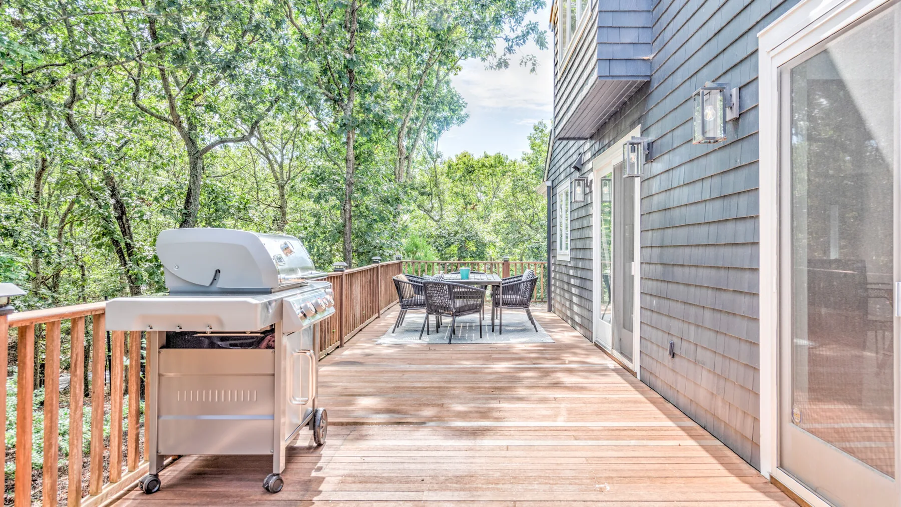 Una terraza de madera junto a una casa tiene una parrilla y un comedor al aire libre.
