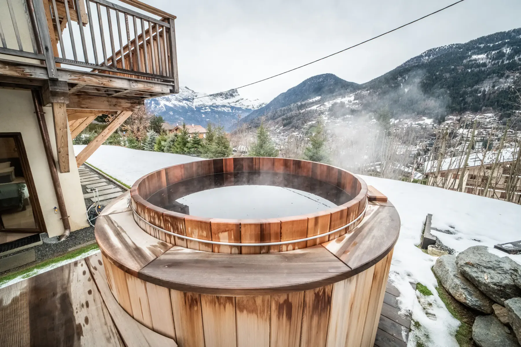 A wooden hot tub sits on an outdoor deck overlooking a snowy mountain valley.