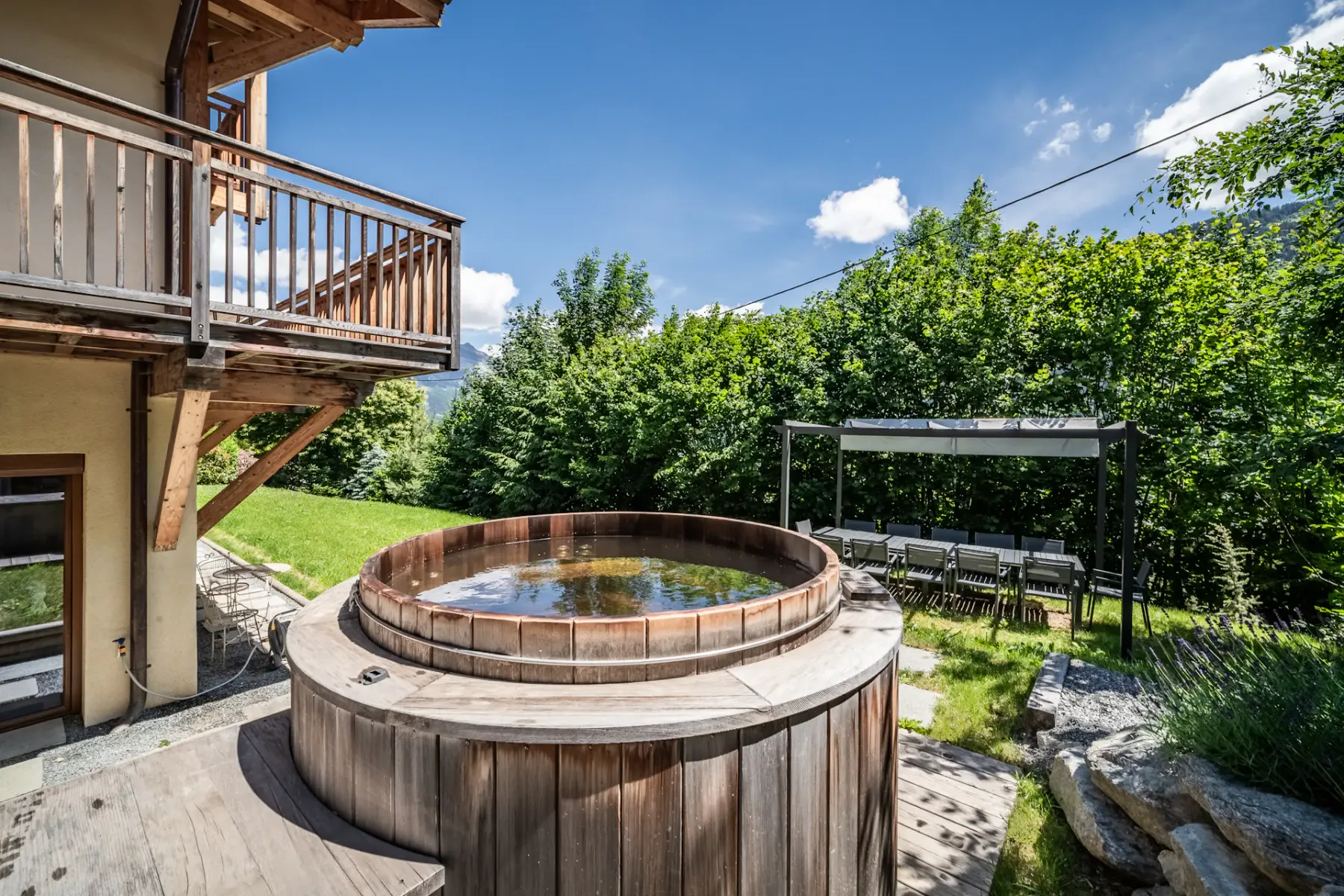 An outdoor hot tub sits beside a chalet, with garden seating and trees nearby.