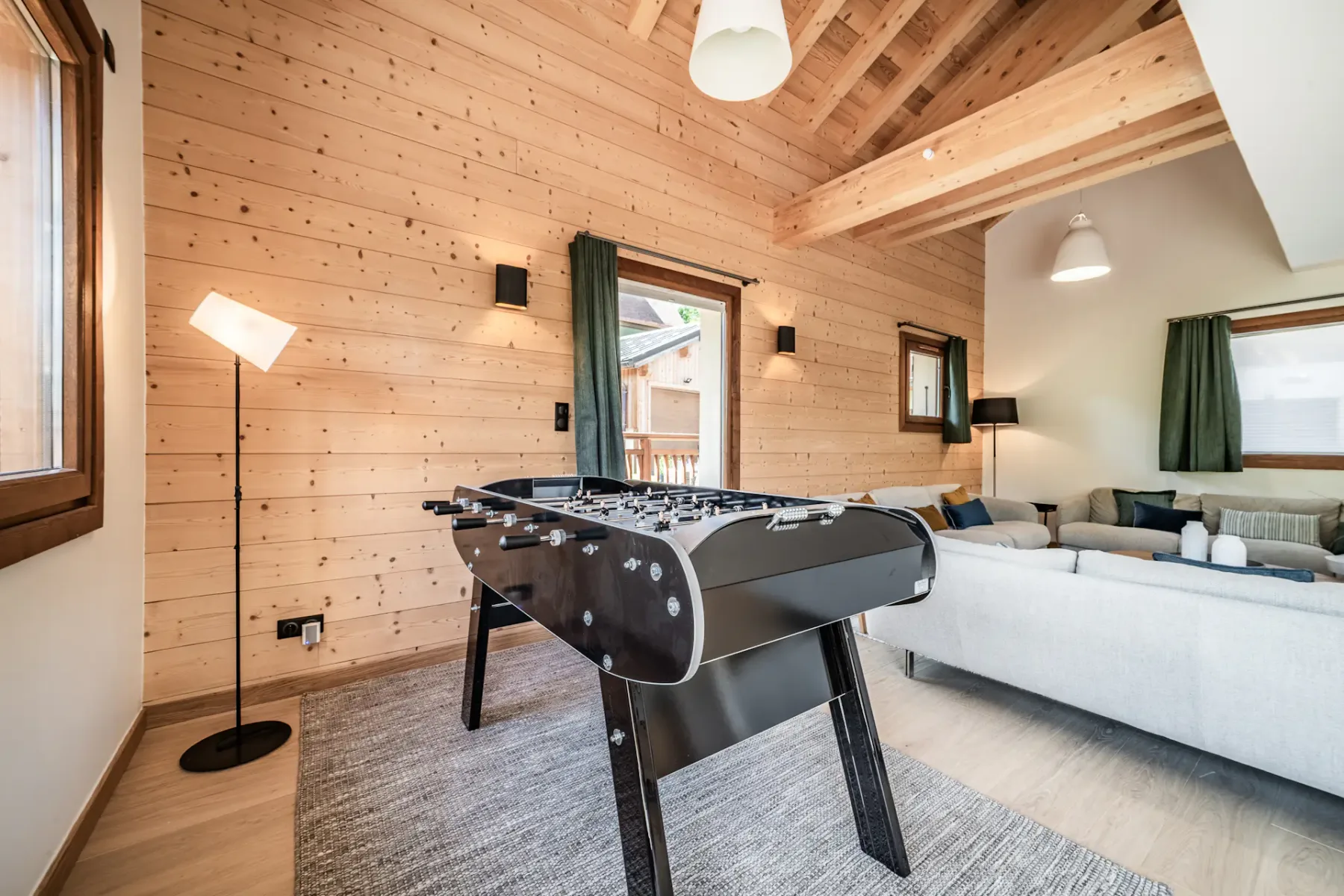 Cozy game room with wooden walls, featuring a foosball table, gray couch, floor lamp, and large windows with green curtains.