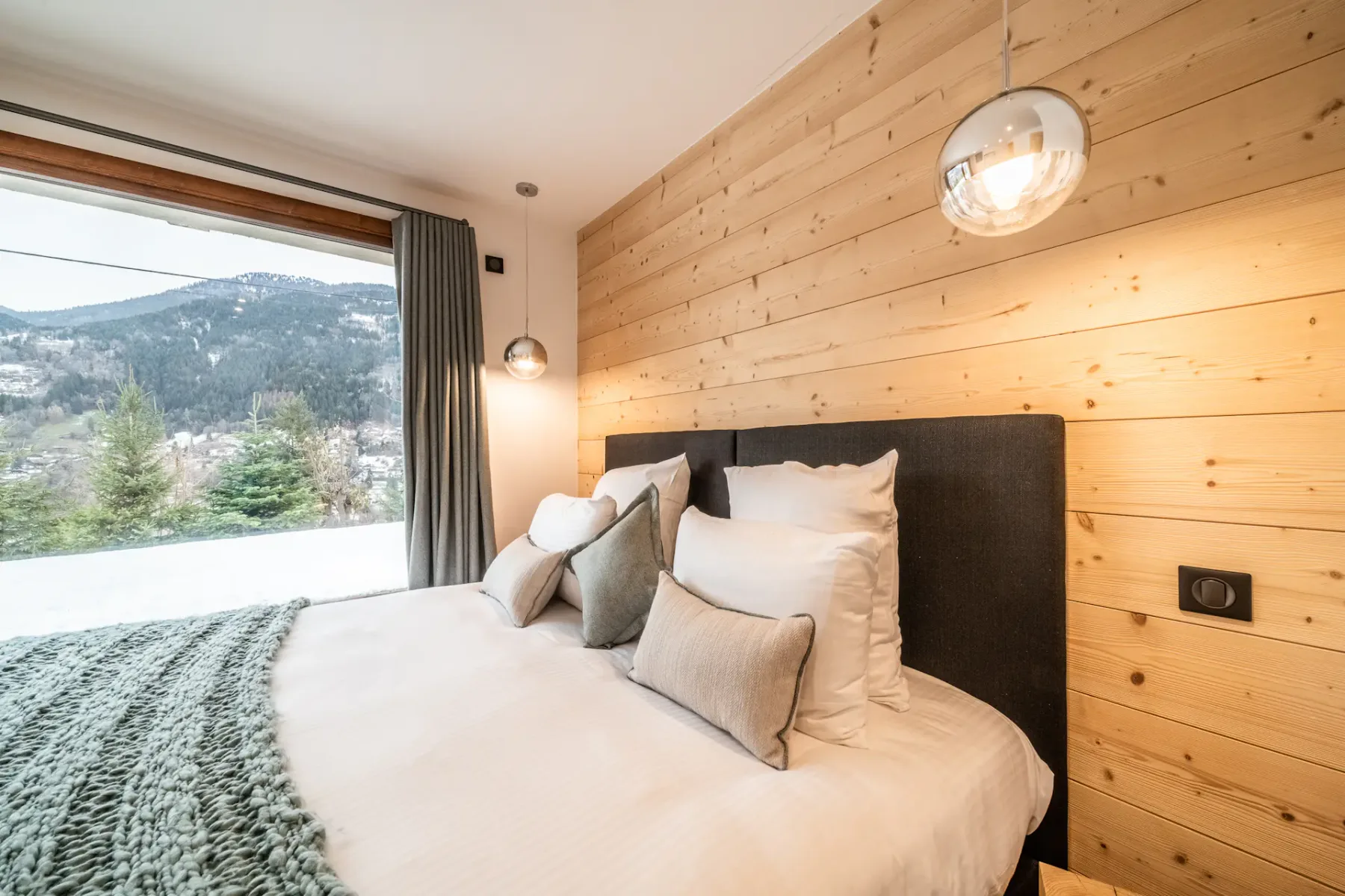 A bedroom with a neatly made bed beside a large window overlooking a mountain landscape.