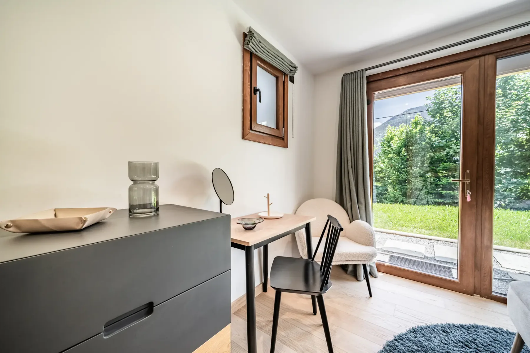 A bright bedroom corner has a desk and seating beside a glass door opening onto greenery.