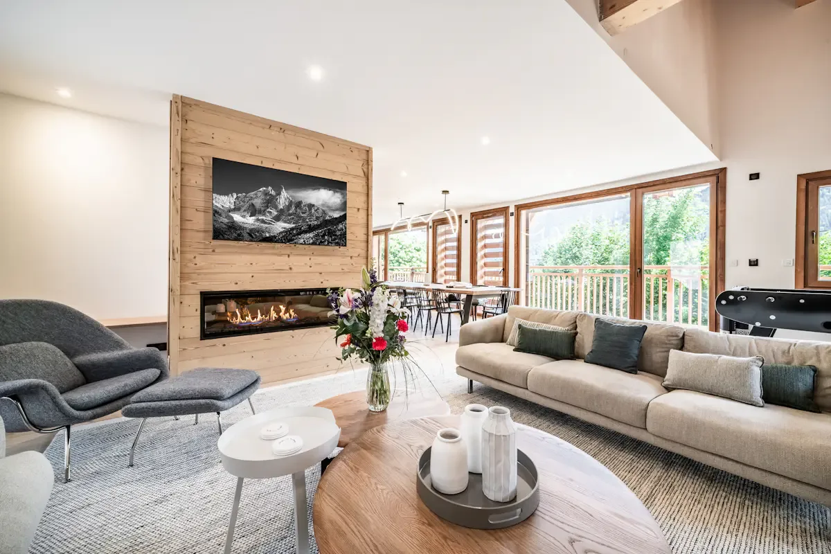 A bright living room has a fireplace, seating, and large windows overlooking greenery.