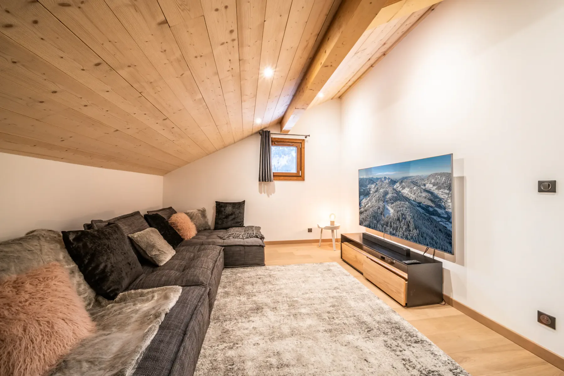 A living room with a sectional sofa, pale rug, and television beneath a sloped wooden ceiling.