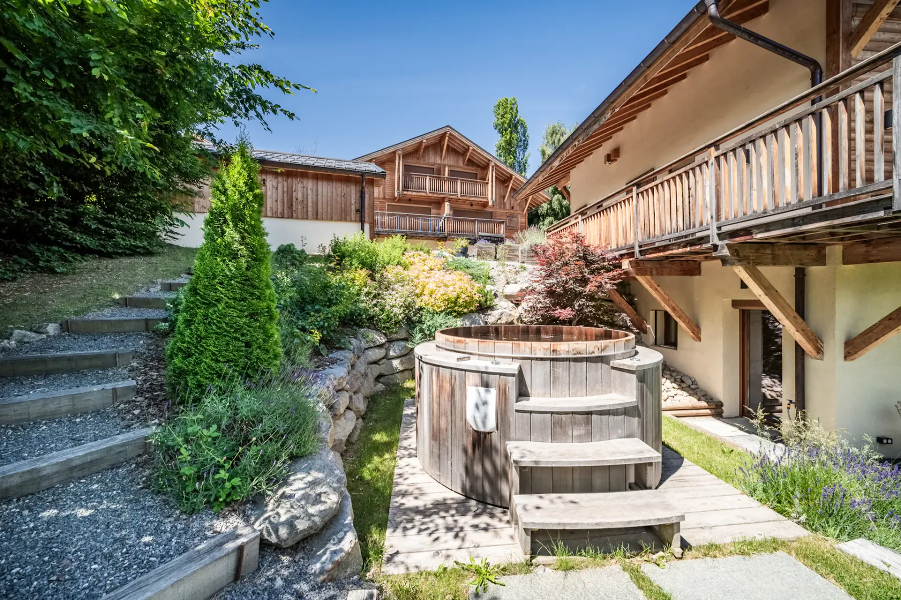 A landscaped outdoor patio features a round hot tub beside a wooden building.