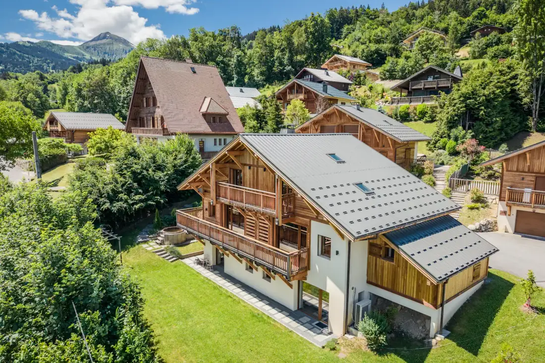 Aerial view of charming wooden chalets surrounded by lush greenery and mountains under a clear blue sky.