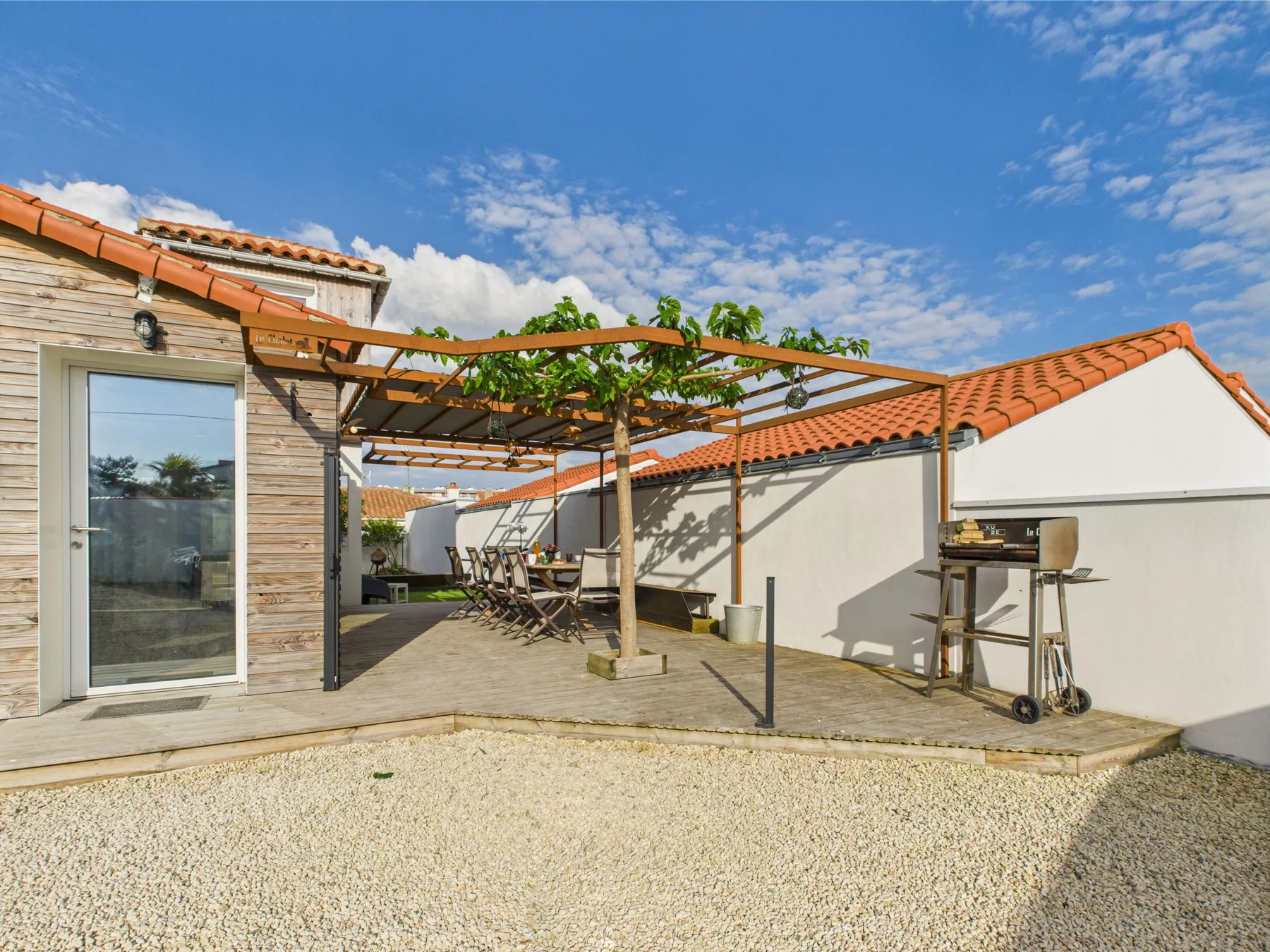 A sunny outdoor patio has a pergola, seating, and a gravel area.