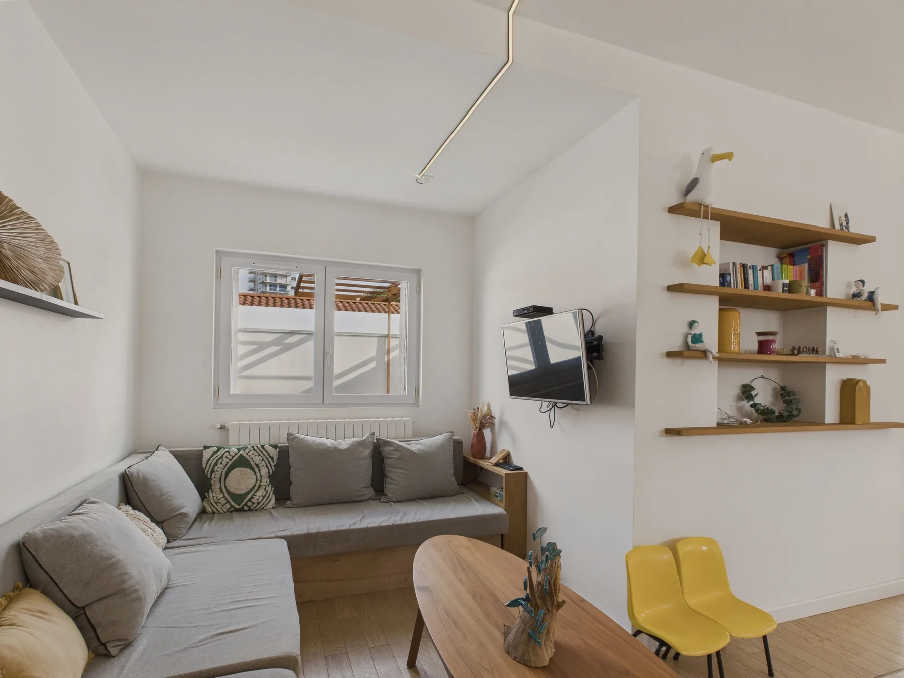 A bright living room with gray seating, a wall-mounted television, and open shelves.