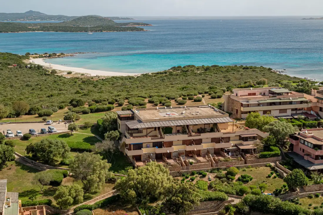 Aerial view of low-rise buildings among greenery beside a beach and turquoise bay.