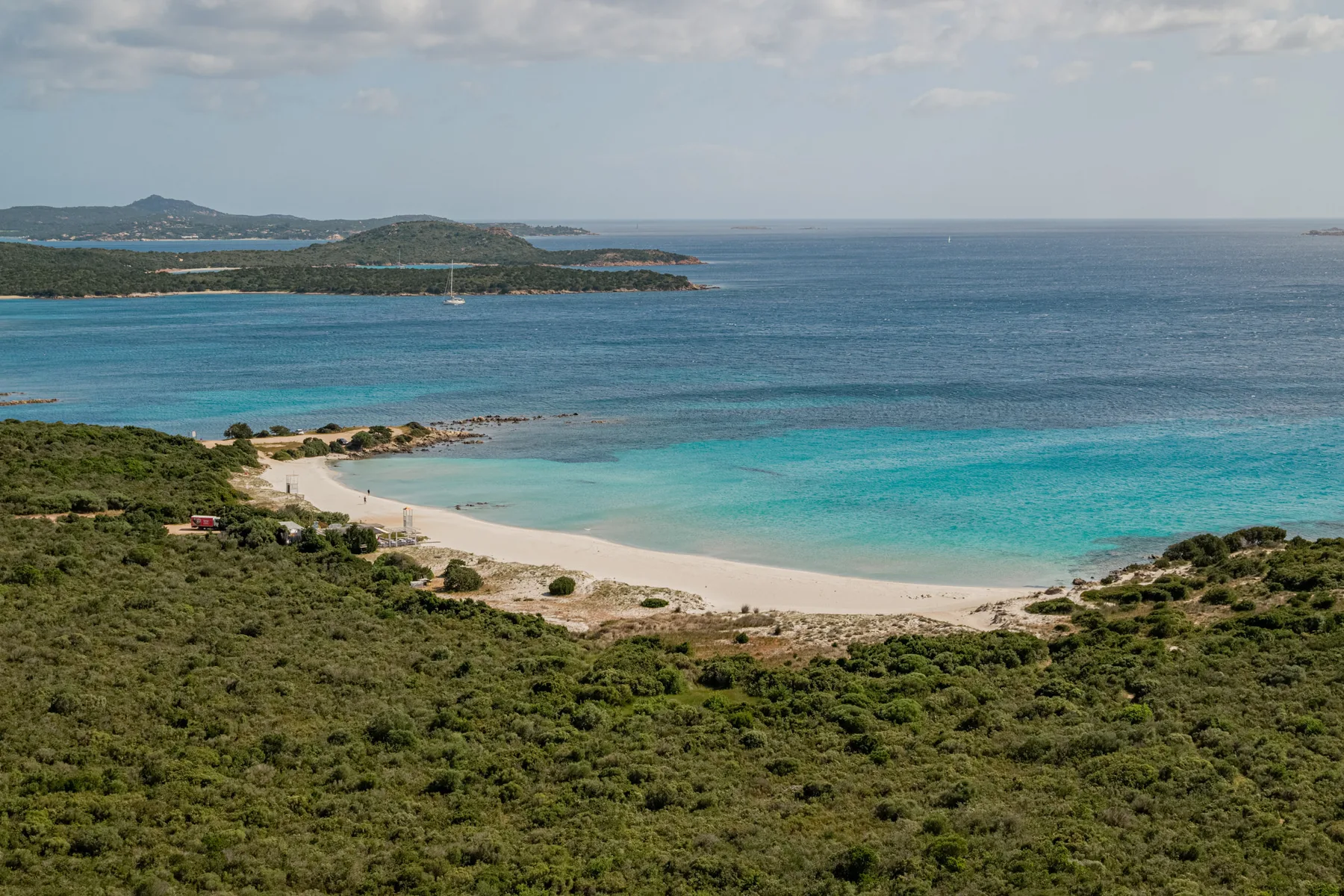 A sandy beach curves along clear turquoise water, with green hills in the foreground.