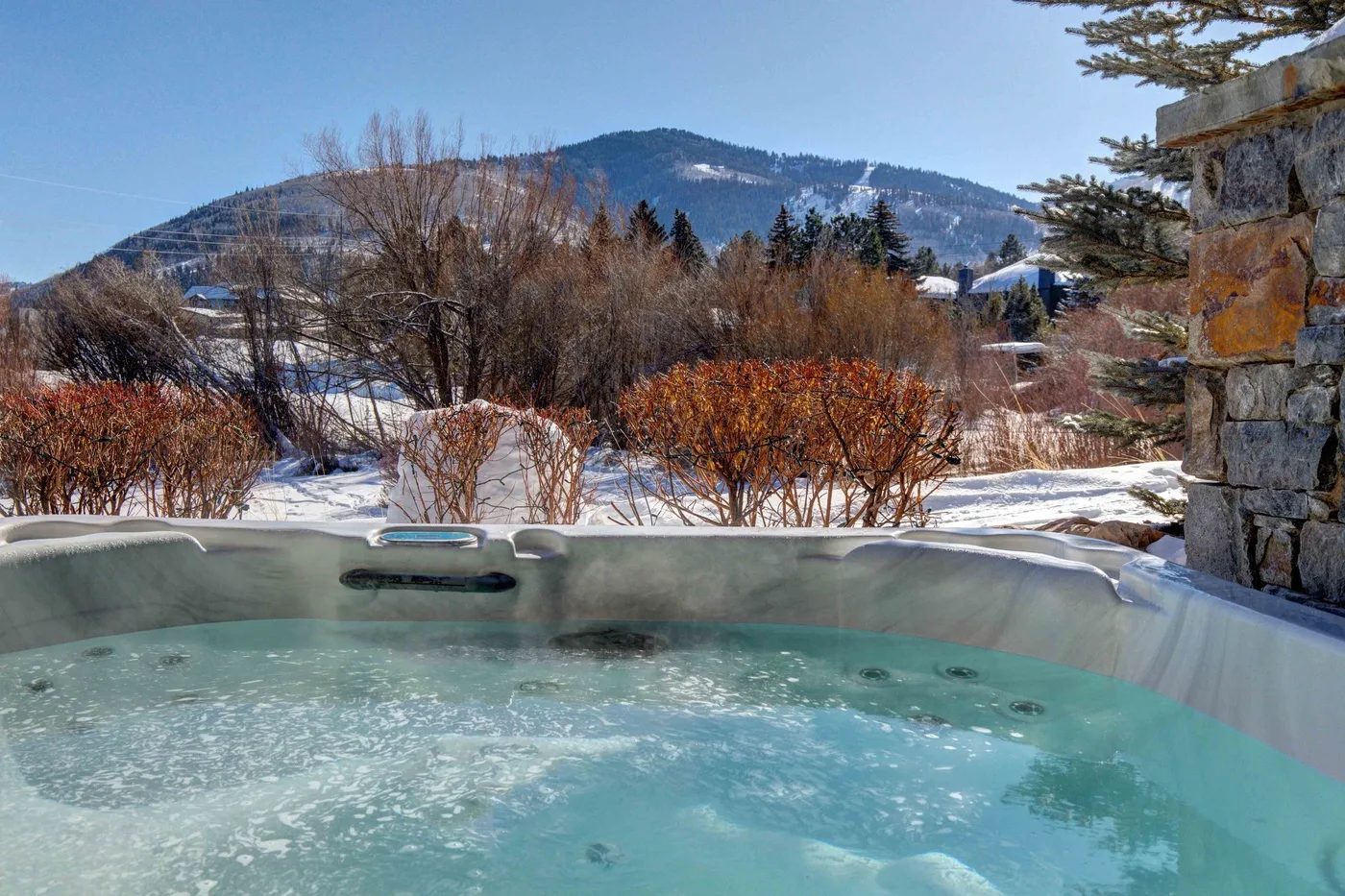 An outdoor hot tub overlooks snow-covered shrubs and mountain scenery.