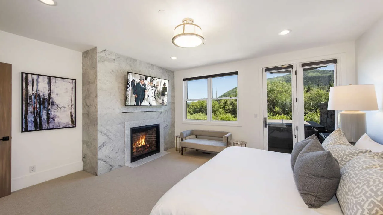 A bright bedroom with a bed, fireplace, and windows overlooking greenery.