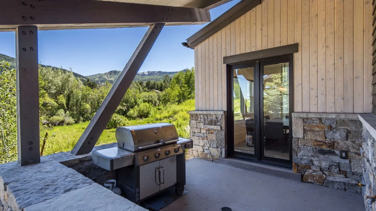 A covered outdoor patio with a grill and sliding glass doors overlooking greenery.