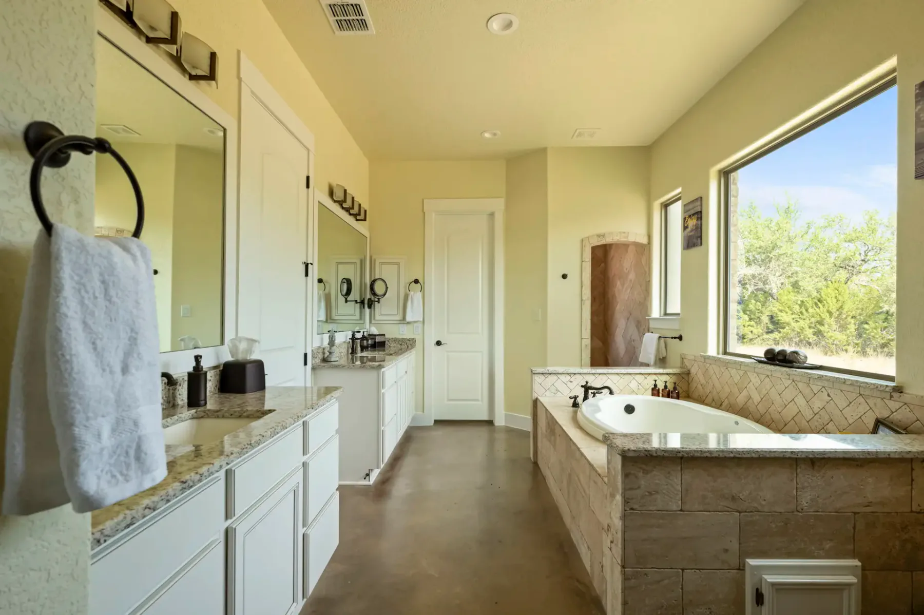 Spacious bathroom with two sinks, granite countertops, a towel rack, elevated bathtub, large window, and wooden floor. Bright natural light.