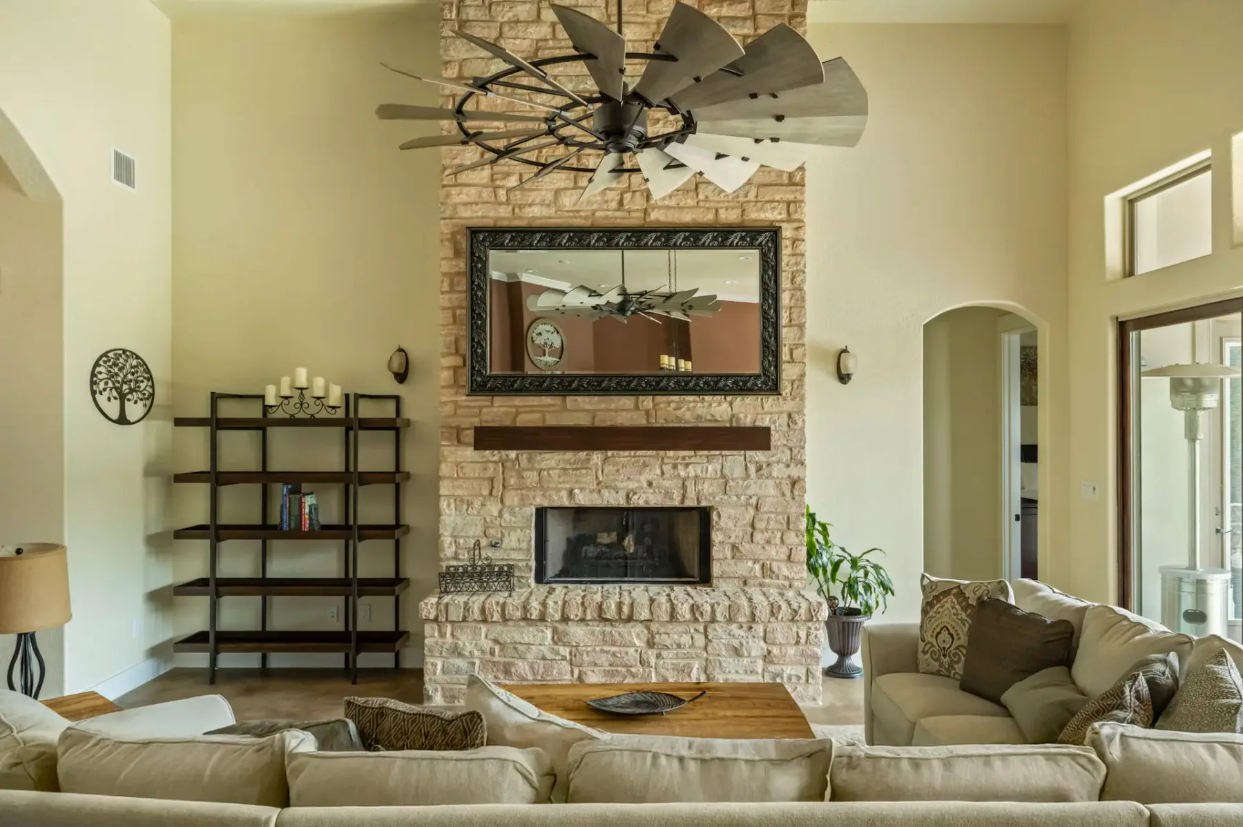 Spacious living room with a stone fireplace, large mirror, beige sectional sofa, wooden coffee table, and metal bookshelf. Neutral decor tones.