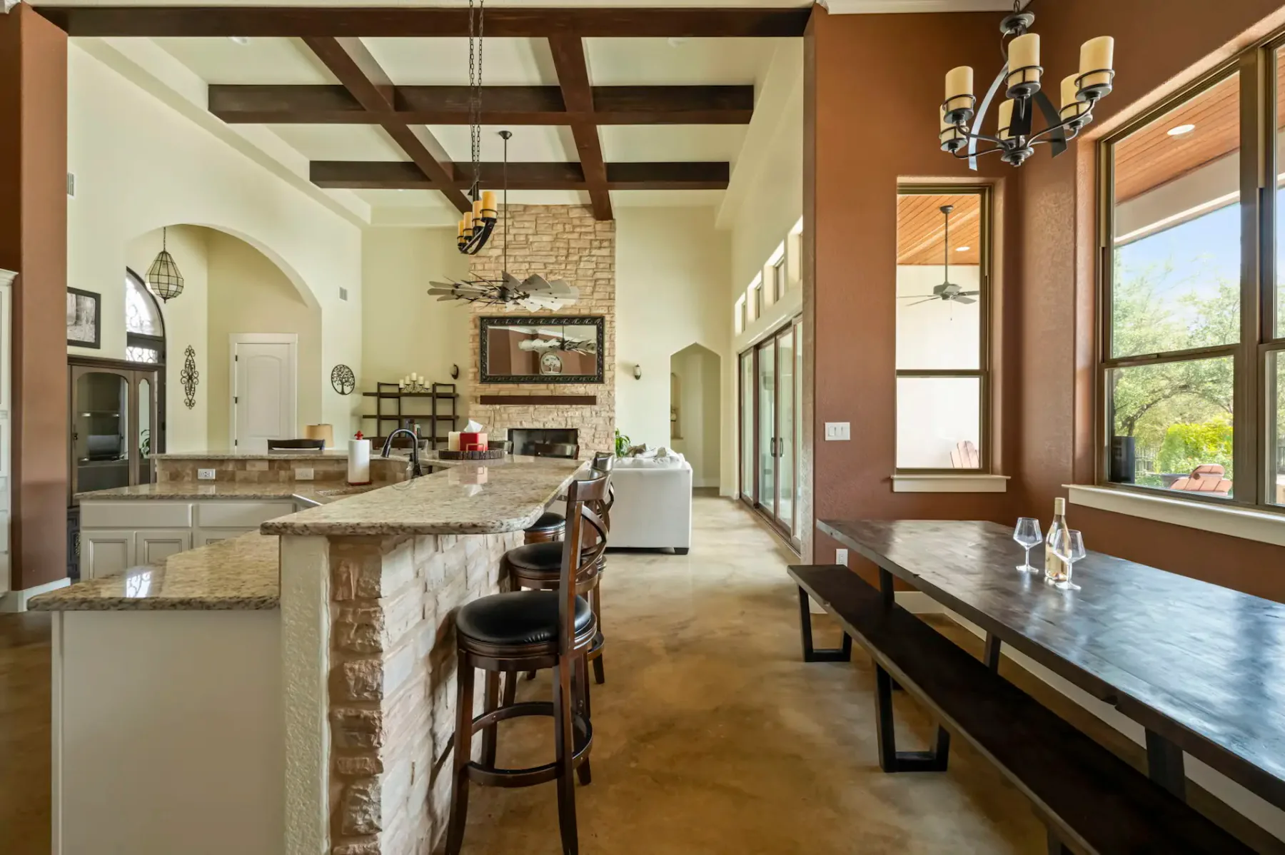 Spacious kitchen with stone island, wooden beams, a rustic dining table, large windows, and a view into the living room with a fireplace.