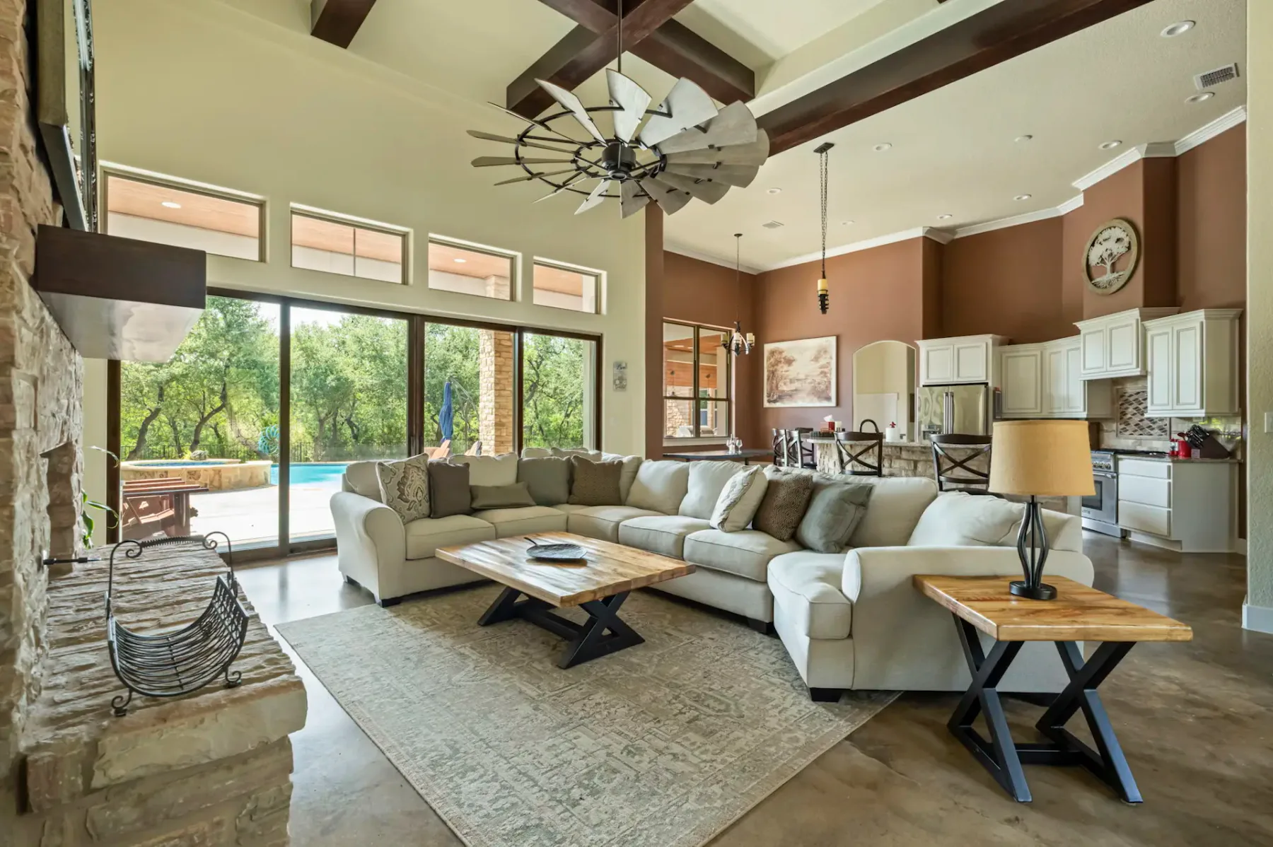 Spacious living room with beige sectional sofa, wooden coffee tables, and large windows overlooking a pool and greenery outside.