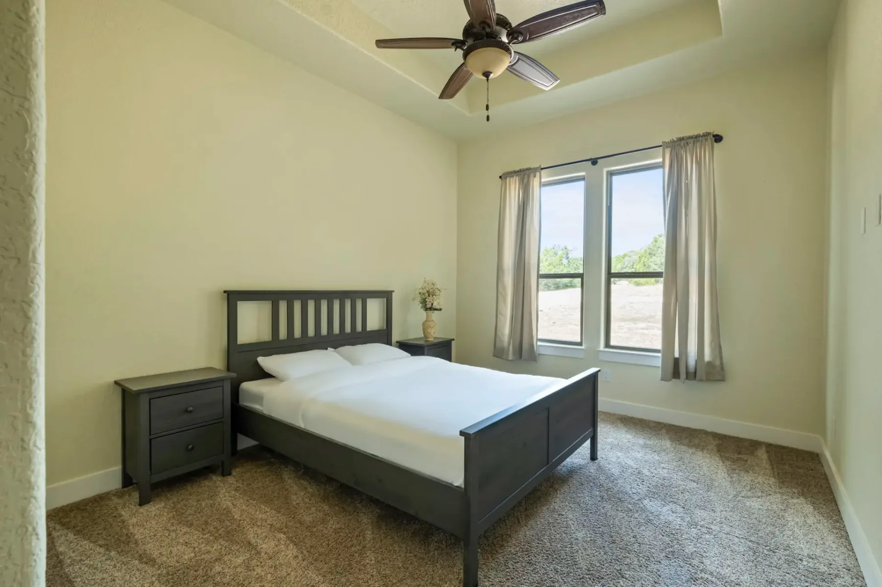 Minimalist bedroom with a dark wood bed, white bedding, matching nightstand, ceiling fan, and large window with sheer curtains overlooking trees.