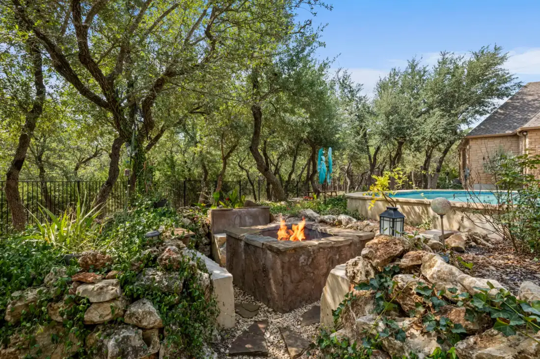 Stone fire pit surrounded by trees and greenery, near a house with a pool, under a clear blue sky.