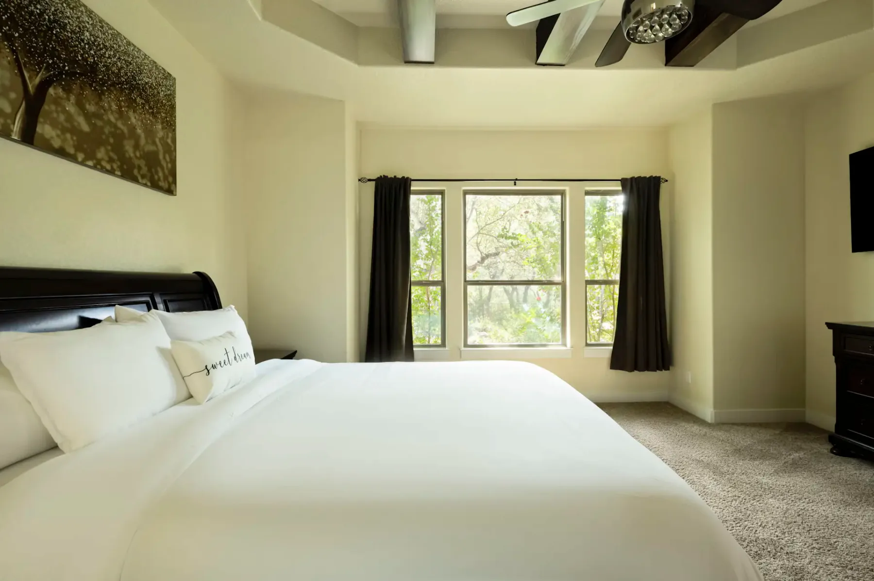 Bright bedroom with a large bed, white bedding, dark curtains, wall art, and a ceiling fan. Sunlight streams through a window with a tree view.
