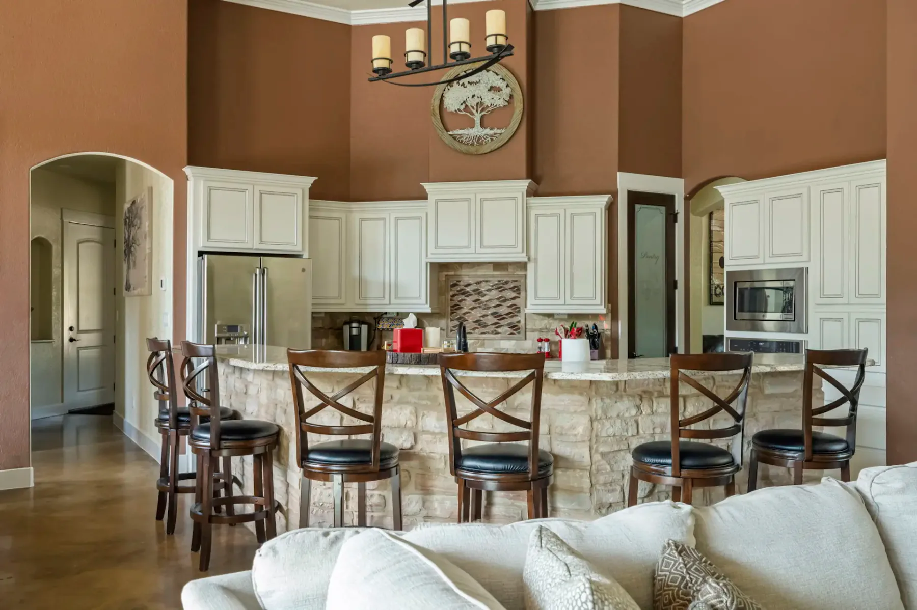Spacious kitchen with brown walls, white cabinets, stone island with chairs, and modern appliances, under a chandelier. Beige couch in foreground.