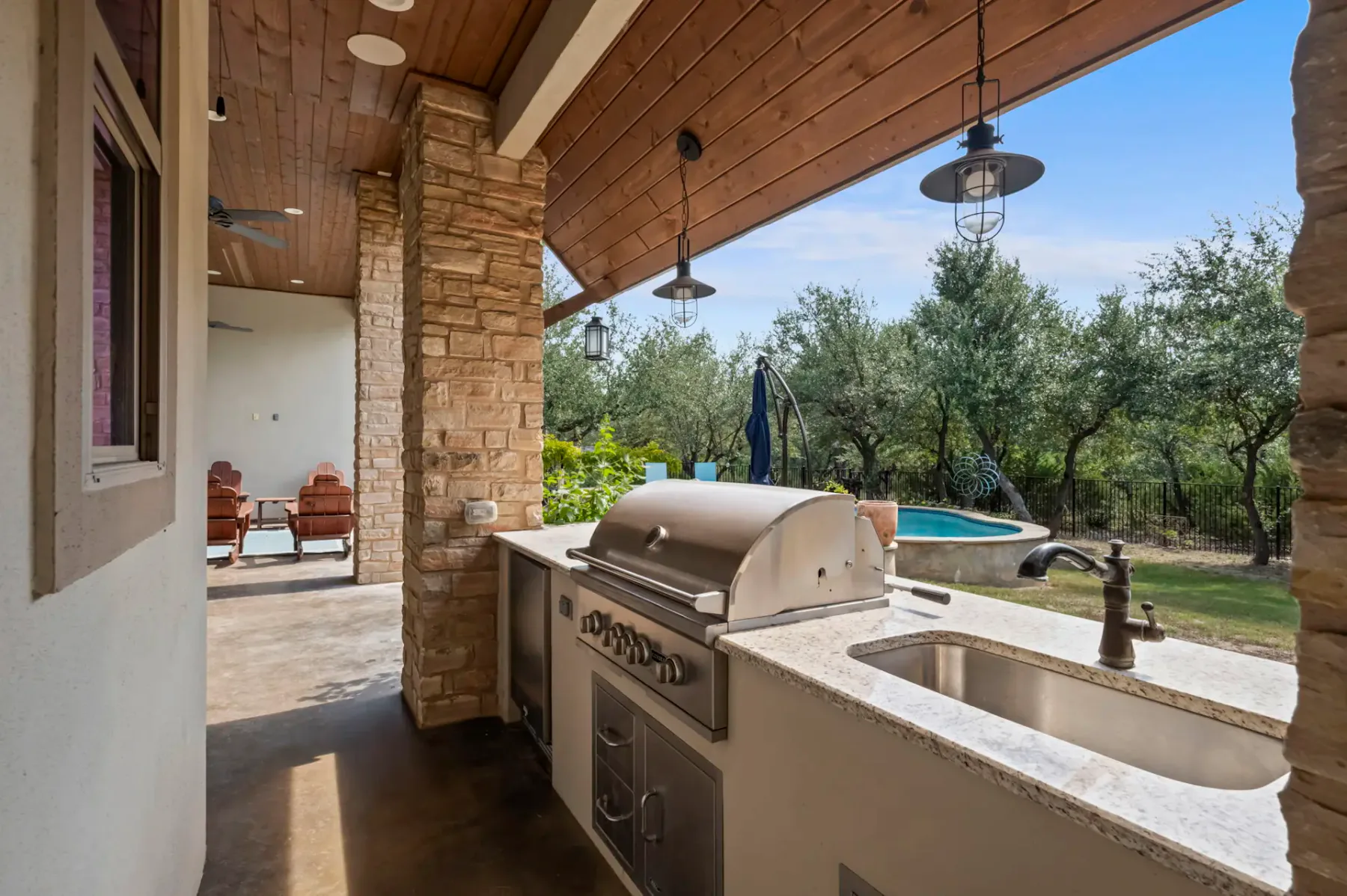 Outdoor kitchen with stone counters, grill, and sink under a wooden roof. Patio chairs and trees visible in the background.