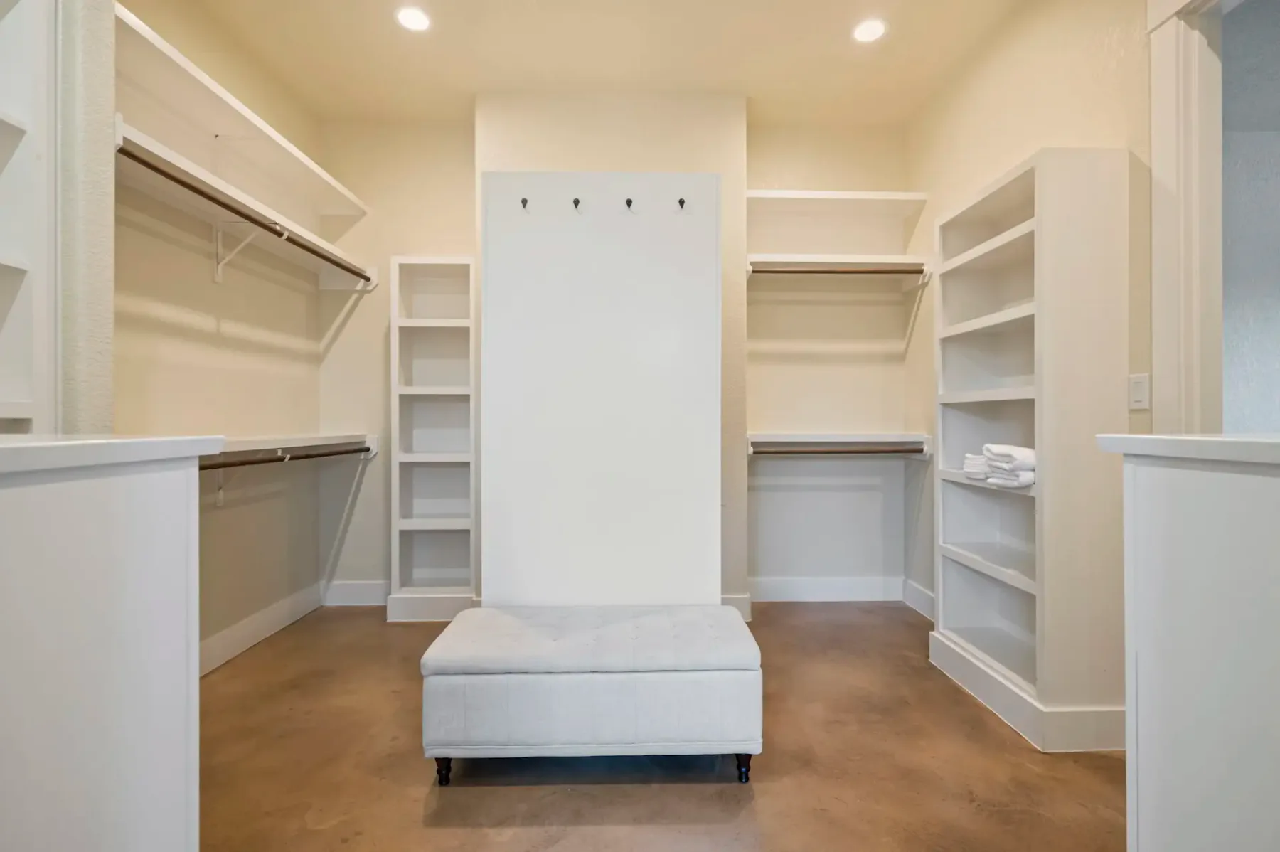 Spacious walk-in closet with white shelves, hanging rods, hooks, and a central upholstered bench on a wooden floor.