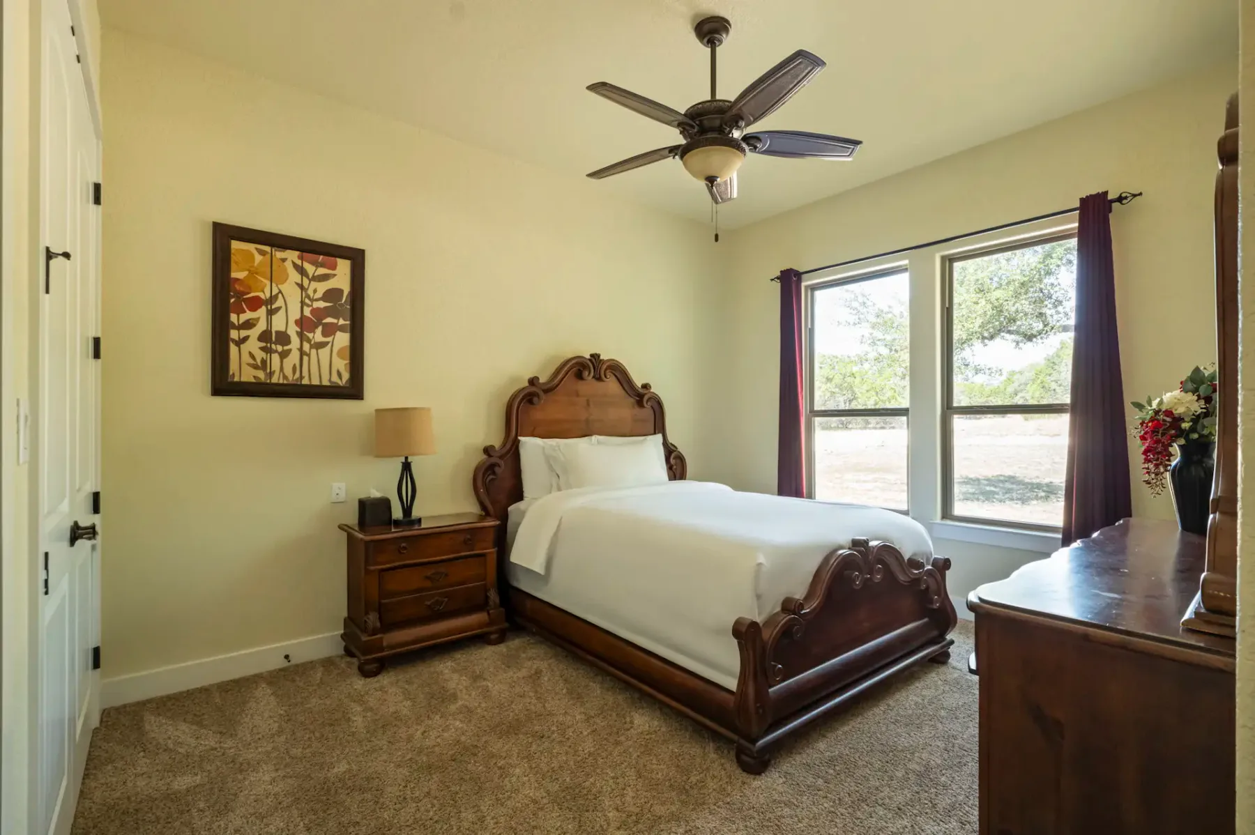 Cozy bedroom with a wooden bed, white bedding, wooden nightstand, lamp, artwork on wall, ceiling fan, and window with red curtains.