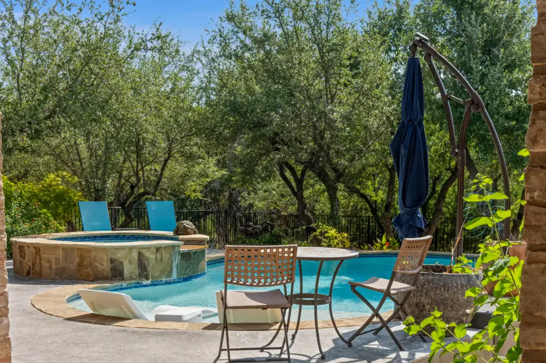 Backyard pool with a stone hot tub, surrounded by trees and a patio with chairs, a table, and a closed umbrella on a sunny day.
