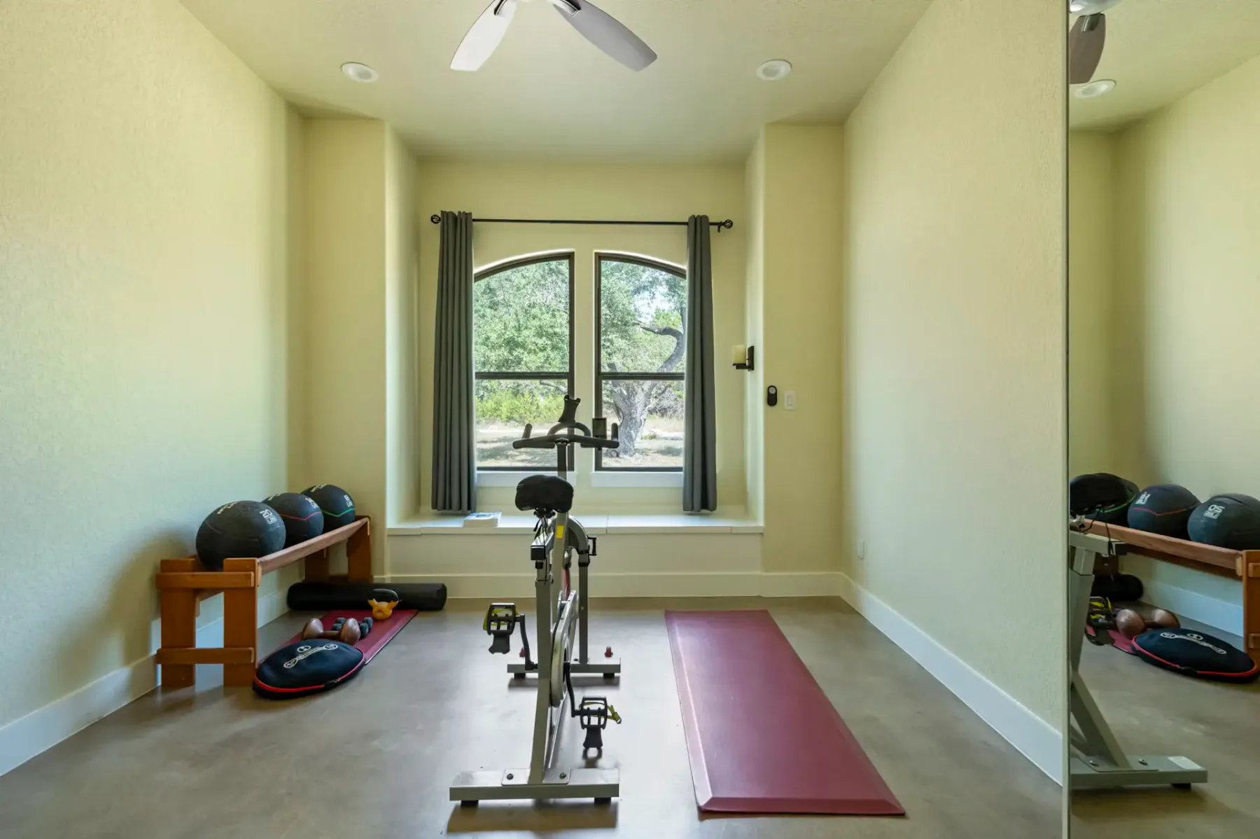 Home gym with an exercise bike, yoga mat, and medicine balls. Large window with curtains and a ceiling fan overhead.