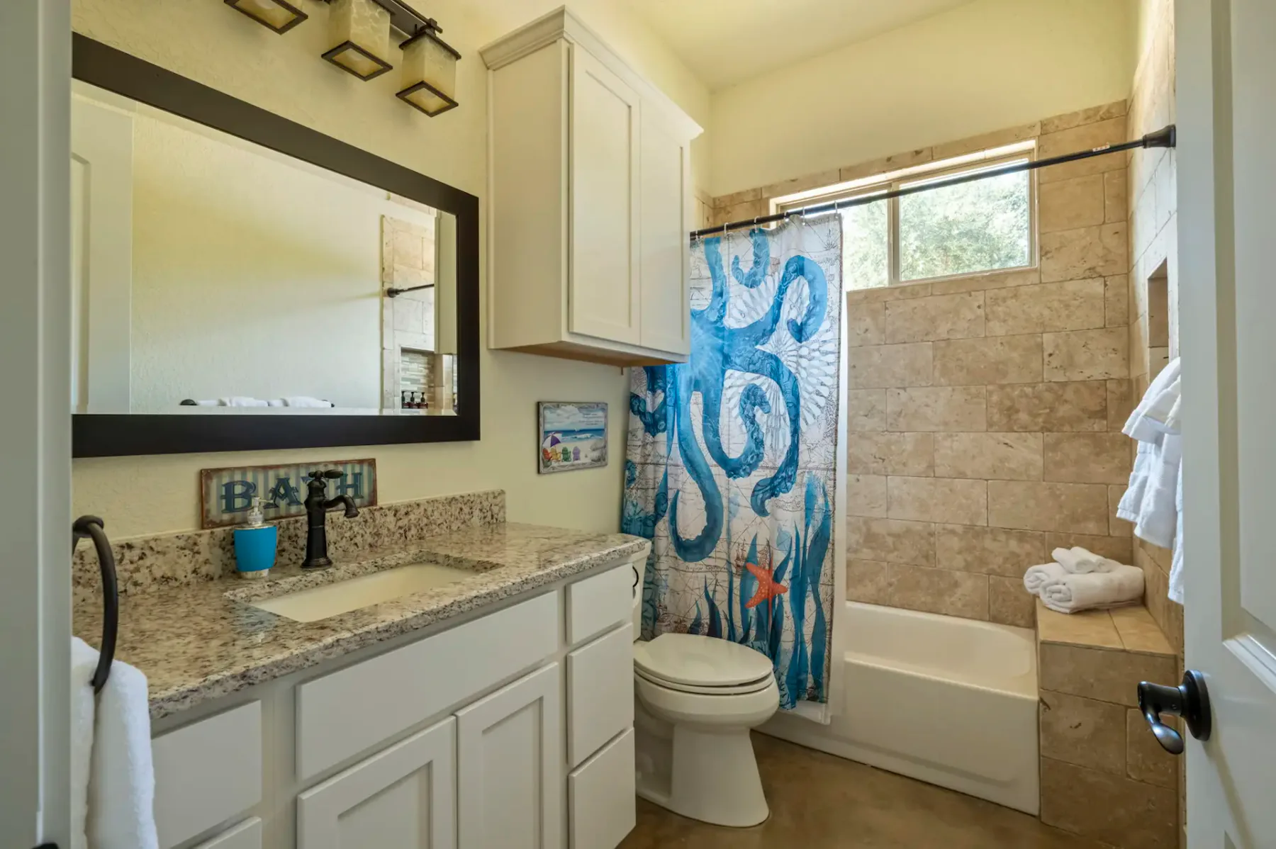 Bathroom with granite countertop, large mirror, white cabinets, shower with ocean-themed curtain, and towel on tub ledge. Natural light from window.