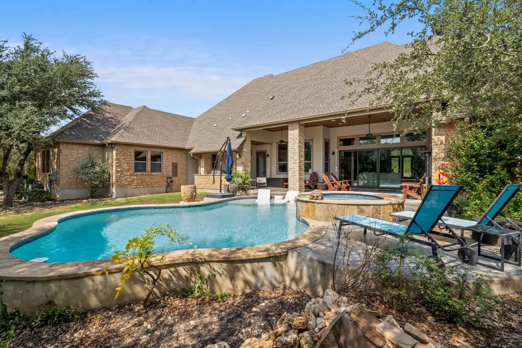 Backyard view of a large house with a stone patio and a curved swimming pool, surrounded by trees and lounge chairs under a clear sky.