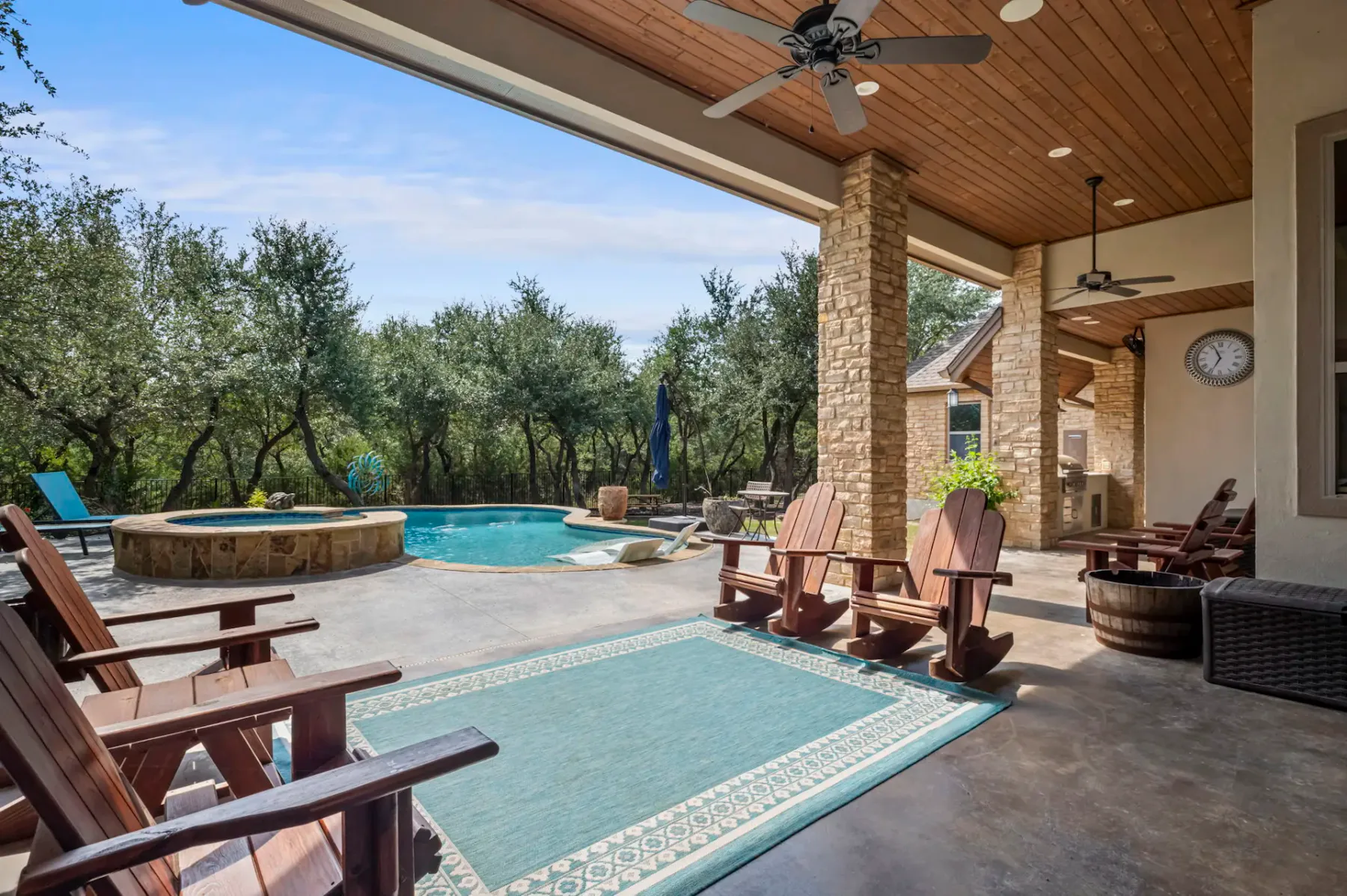 Outdoor patio with wooden chairs, a rug, and ceiling fans overlooks a pool and hot tub surrounded by trees and stone pillars.