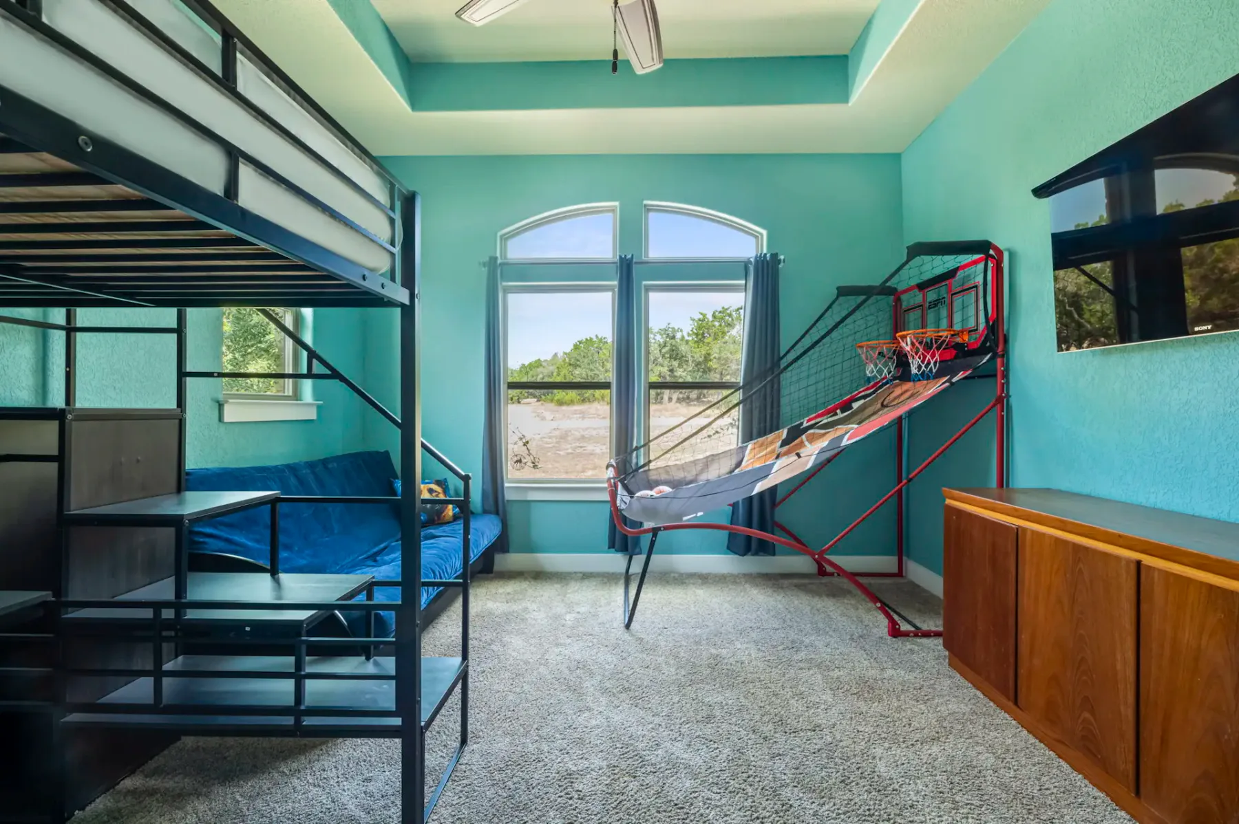 Kids' room with a bunk bed, blue walls, large windows, a basketball hoop game, a blue couch, and a wooden cabinet. Carpeted floor.